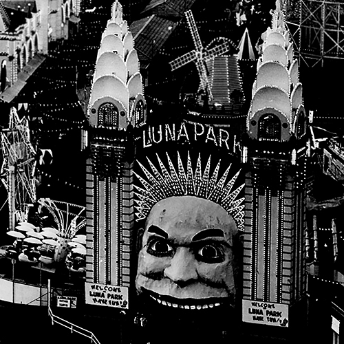 Historical photograph of the iconic entrance to Sydney Luna Park. Two Art Deco spires rise either side of the entrance, a giant garish smiling face. Glittering lights edge every contour, creating a sparkling array.